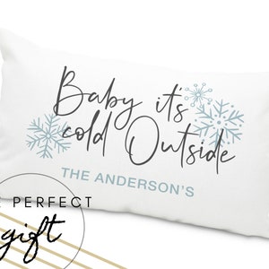 May include: White throw pillow with the text "Baby it's cold outside" in black cursive font. The text "THE ANDERSON'S" is below the main text. The pillow has a blue snowflake design.