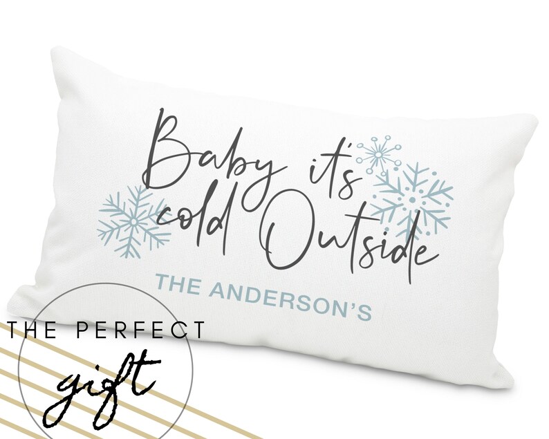May include: White throw pillow with the text "Baby it's cold outside" in black cursive font. The text "THE ANDERSON'S" is below the main text. The pillow has a blue snowflake design.