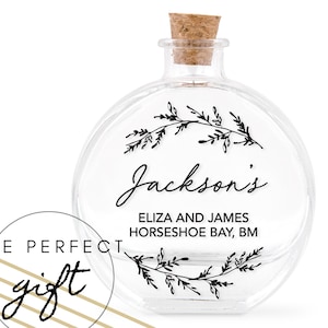 Op de afbeelding: Een heldere, ronde glazen fles met een kurk. De fles is versierd met zwarte bloemmotieven en de naam "Jackson's" in elegant schrift. Extra tekst is "ELIZA AND JAMES HORSESHOE BAY, BM". Een cirkelvormige afbeelding leest "THE PERFECT gift".