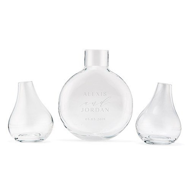 May include: Three clear glass vases. The central vase is round with a short neck and features the names "Alexis and Jordan" and the date "05.05.2019". Two smaller, teardrop-shaped vases flank the central vase.