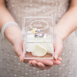 May include: A clear acrylic ring box with a heart and the initials "ee" engraved on the lid. The box is filled with white rose petals and contains a silver engagement ring.
