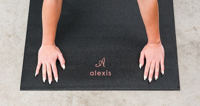 May include: A black rectangular yoga mat with a textured surface. The mat features a personalised design in the centre with a large cursive letter A above the name alexis, both printed in a metallic rose gold colour on the dark mat surface.