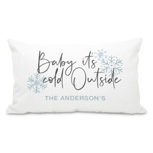 May include: White throw pillow with the text "Baby it's cold outside" in black script font and blue snowflake designs. The text "THE ANDERSON'S" is printed below the main text.