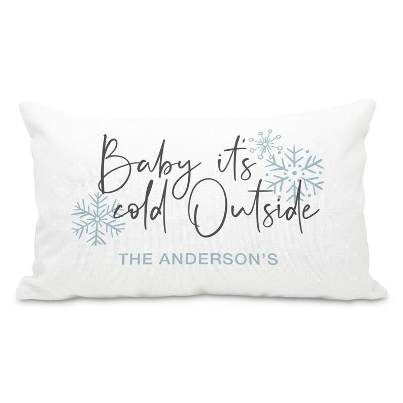 May include: White throw pillow with the text "Baby it's cold outside" in black script font and blue snowflake designs. The text "THE ANDERSON'S" is printed below the main text.