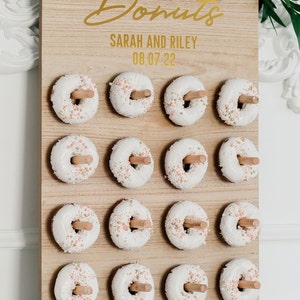 May include: A wooden donut wall with the text "Donuts" in gold script, "Sarah and Riley" and "08.07.22" in gold lettering. The wall is decorated with 16 white donuts with pink and gold sprinkles.