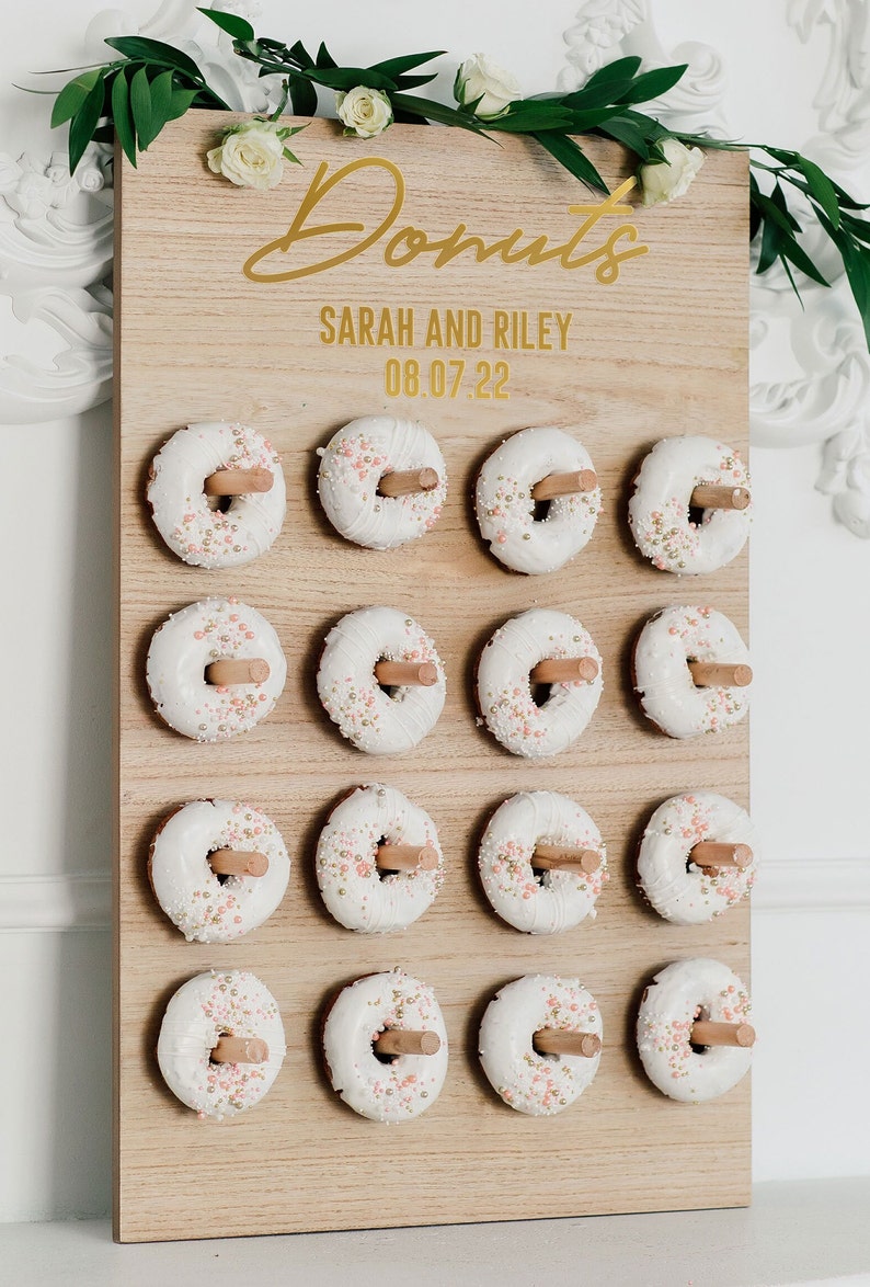May include: A wooden donut wall with the text "Donuts" in gold script, "Sarah and Riley" and "08.07.22" in gold lettering. The wall is decorated with 16 white donuts with pink and gold sprinkles.