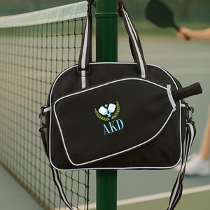 Könnte beinhalten: Schwarze Pickleball-Tasche mit weißen Zierleisten und Griffen, die an einer grünen Stange in der Nähe eines Tennisnetzes hängt. Die Tasche hat einen passenden Pickleball-Schläger und ein Logo mit den Initialen "AKD". Im Hintergrund spielt eine Person Pickleball.
