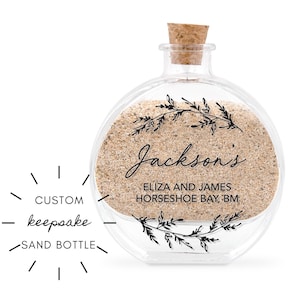 May include: A clear, round glass sand bottle with a cork stopper. The bottle is filled with sand and features the text "Jackson's" and "ELIZA AND JAMES HORSESHOE BAY, BM". Decorative floral elements are printed on the sand. The text "CUSTOM keepsake SAND BOTTLE" is also present.