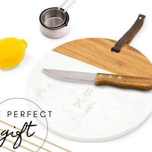 May include: A round white and brown chopping board with a knife and a leather handle. The board has a monogram of the letters "B" and "K" with a small branch design.