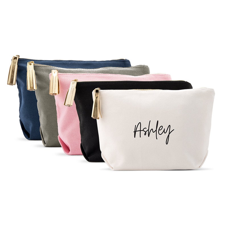 May include: Five canvas makeup bags with gold tassels. The bags are in navy blue, gray, pink, black, and white. The white bag has the name "Ashley" written on it in black.