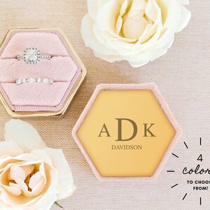 May include: A hexagon-shaped ring box with a pink velvet interior. The box is open and contains two rings. The box is gold with a pink border. The box is sitting on a beige surface. The text "ADK DAVIDSON" is printed on the box. The text "4 colors to choose from!" is printed on the surface.