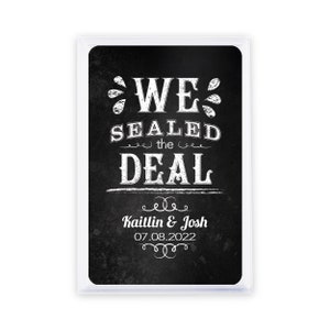 May include: A rectangular sign with a black chalkboard background and white text that reads "WE SEALED THE DEAL". Below the text are the names "Kaitlin & Josh" and the date "07.08.2022". The sign is encased in a clear plastic frame.