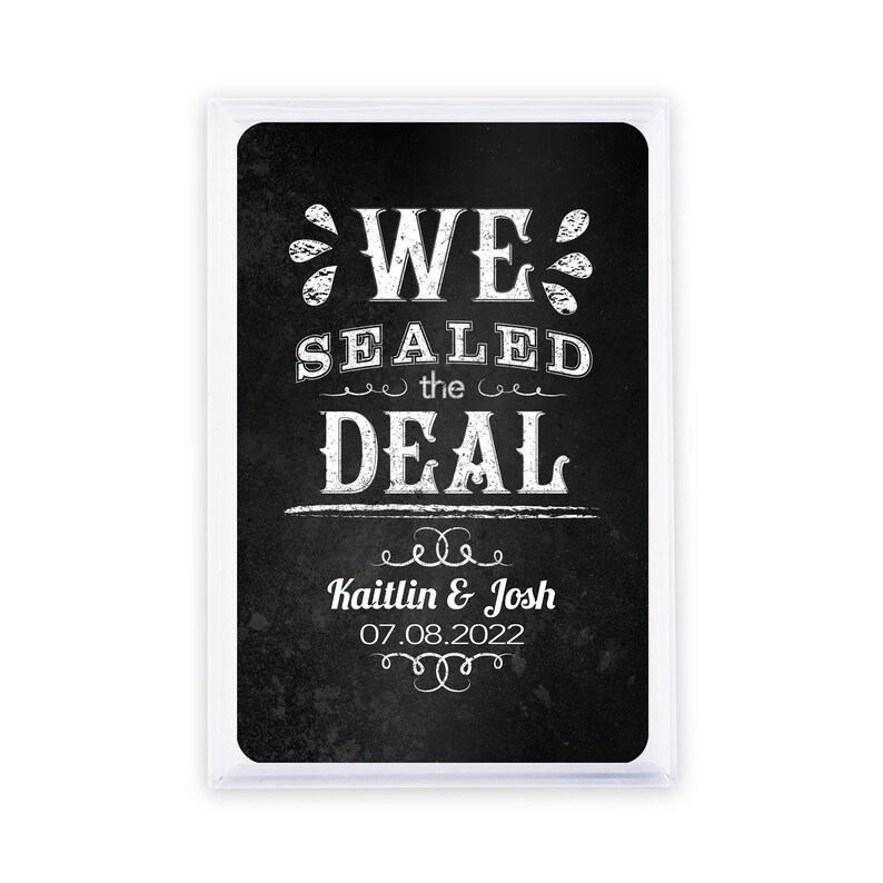 May include: A rectangular sign with a black chalkboard background and white text that reads "WE SEALED THE DEAL". Below the text are the names "Kaitlin & Josh" and the date "07.08.2022". The sign is encased in a clear plastic frame.