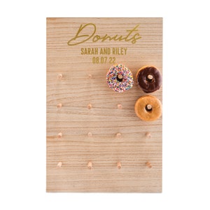 May include: A wooden donut wall with the text "Donuts" in gold script, "Sarah and Riley" and "08.07.22" in gold text. The wall has holes for donuts and three donuts are displayed: one with sprinkles, one with chocolate frosting, and one with plain glaze.