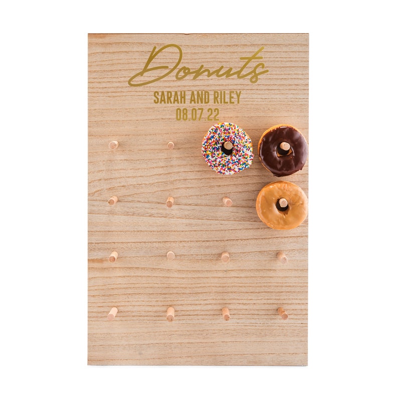 May include: A wooden donut wall with the text "Donuts" in gold script, "Sarah and Riley" and "08.07.22" in gold text. The wall has holes for donuts and three donuts are displayed: one with sprinkles, one with chocolate frosting, and one with plain glaze.