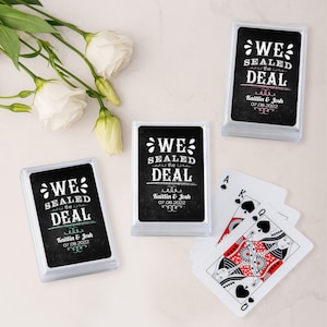 May include: Clear plastic cases containing playing cards with a black background and white text that reads "WE SEALED THE DEAL" with names and a date. A few playing cards are fanned out next to the cases. White flowers are in the background.