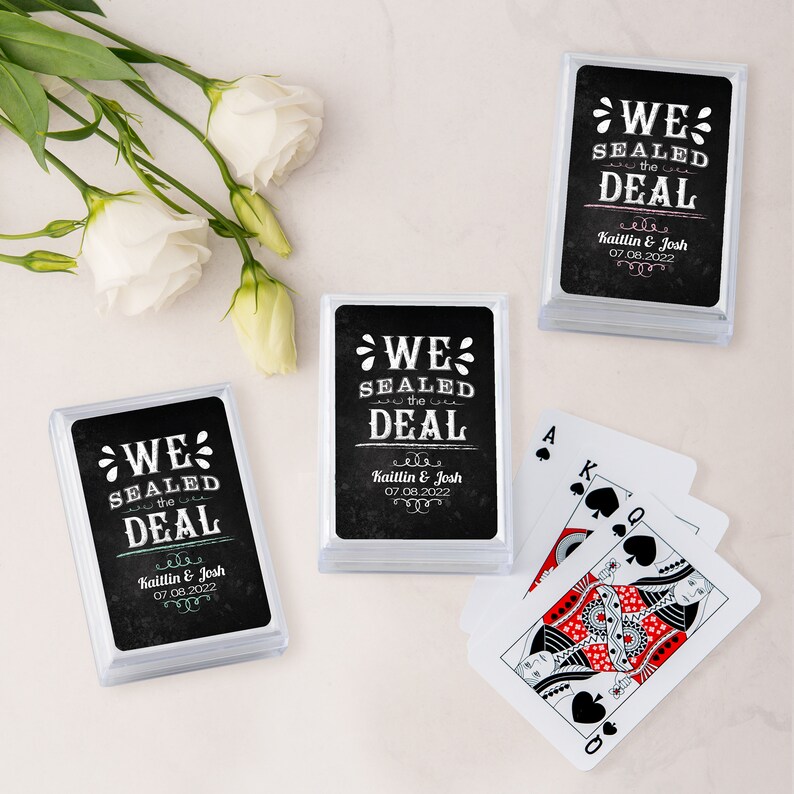 May include: Clear plastic cases containing playing cards with a black background and white text that reads "WE SEALED THE DEAL" with names and a date. A few playing cards are fanned out next to the cases. White flowers are in the background.