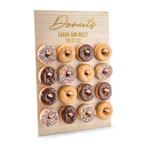 May include: A wooden donut wall with the text "Donuts" in gold script, "Sarah and Riley" and "08.07.22" in gold lettering. The wall is decorated with 16 donuts in various flavors, including chocolate, sprinkles, and plain glazed.
