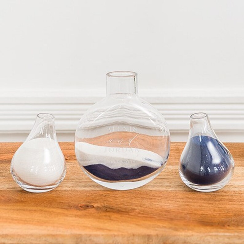 May include: Three clear glass vessels filled with colored sand. The largest is round, the others are teardrop-shaped. The sand colors are white, tan, and navy blue. The largest vessel has text that reads "Alexis and Jordan".