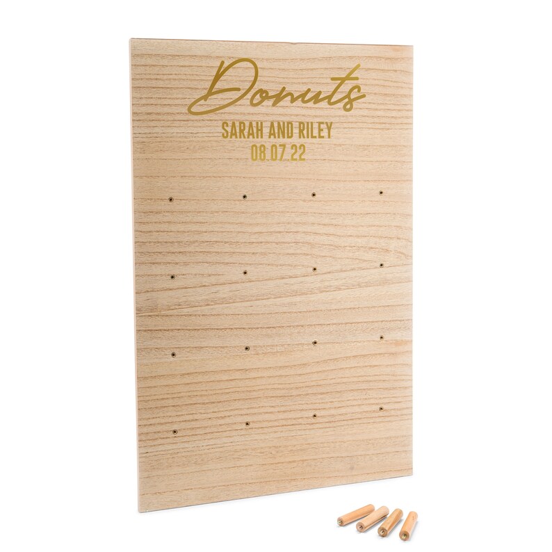 May include: A wooden donut wall with holes for donuts. The wall is light brown with a gold inscription that reads "Donuts, Sarah and Riley, 08.07.22". The wall is accompanied by five wooden dowels.