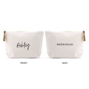 May include: A white canvas makeup bag with a gold zipper and tassel. The front of the bag says "Ashley" in black script. The back of the bag says "BRIDESMAID" in black.