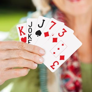 May include: A hand holding playing cards, including a King of Hearts, a Joker, a Jack of Spades, and two cards with the number 3 and the suit of diamonds. The cards are white with red and black symbols.