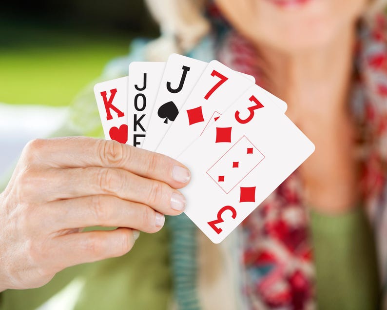 May include: A hand holding playing cards, including a King of Hearts, a Joker, a Jack of Spades, and two cards with the number 3 and the suit of diamonds. The cards are white with red and black symbols.