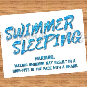 May include: White rectangular sign with the words "SWIMMER SLEEPING" in blue, water-textured font. Below, the text reads "WARNING: WAKING SWIMMER MAY RESULT IN A HIGH-FIVE IN THE FACE WITH A SHARK."
