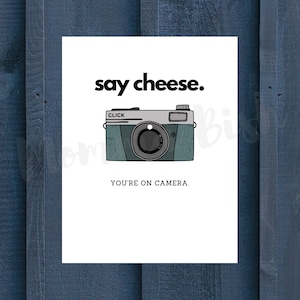 May include: A white card with the text "say cheese." and a cartoon illustration of a vintage camera with the text "CLICK" on the top. Below the camera is the text "YOU'RE ON CAMERA."