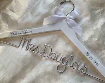 Könnte beinhalten: Ein weißer Holzkleiderbügel mit einem silbernen Drahtschriftzug, der Mrs. Douglas liest. Der Bügel ist oben mit einer weißen Satinschleife verziert und enthält den aufgedruckten Text Chase and Lauren sowie May 20, 1993.