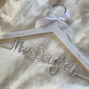 May include: A white wooden wedding dress hanger with a silver hook and a white satin bow. The hanger is personalised with the names "Chase and Lauren" and the date "May 20, 1993". The name "Mrs. Douglas" is crafted in silver wire.