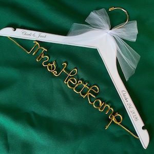May include: A white wooden clothes hanger with a gold metal hook and a gold wire script reading Mrs. Heithkamp. The hanger features a white tulle bow and black printed text reading Charli & Jacob and October 24, 2026.