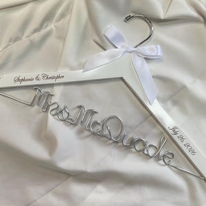 Peut inclure: Un cintre en bois blanc avec un crochet en métal argenté et un nœud en ruban de satin blanc. Le cintre est personnalisé avec le texte Stephanie & Christopher et July 26, 2026, et comporte une inscription en fil métallique Mrs. McQuade.