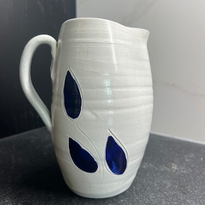 May include: White ceramic pitcher with a blue leaf design. The pitcher has a rounded body and a handle.