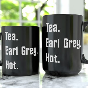 Könnte beinhalten: Zwei schwarze Keramikbecher mit weißem Text, der "Tea. Earl Grey. Hot." lautet.