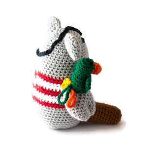 Puede incluir: Un animal de peluche gris tejido a crochet con un parche negro en el ojo y una camisa a rayas rojas y blancas. El animal sostiene un loro verde tejido a crochet con detalles naranjas y amarillos.