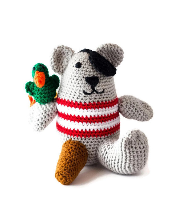 Puede incluir: Un oso de peluche gris tejido a crochet que lleva una camisa a rayas rojas y blancas y un parche negro en el ojo. El oso sostiene un loro tejido a crochet verde y naranja.