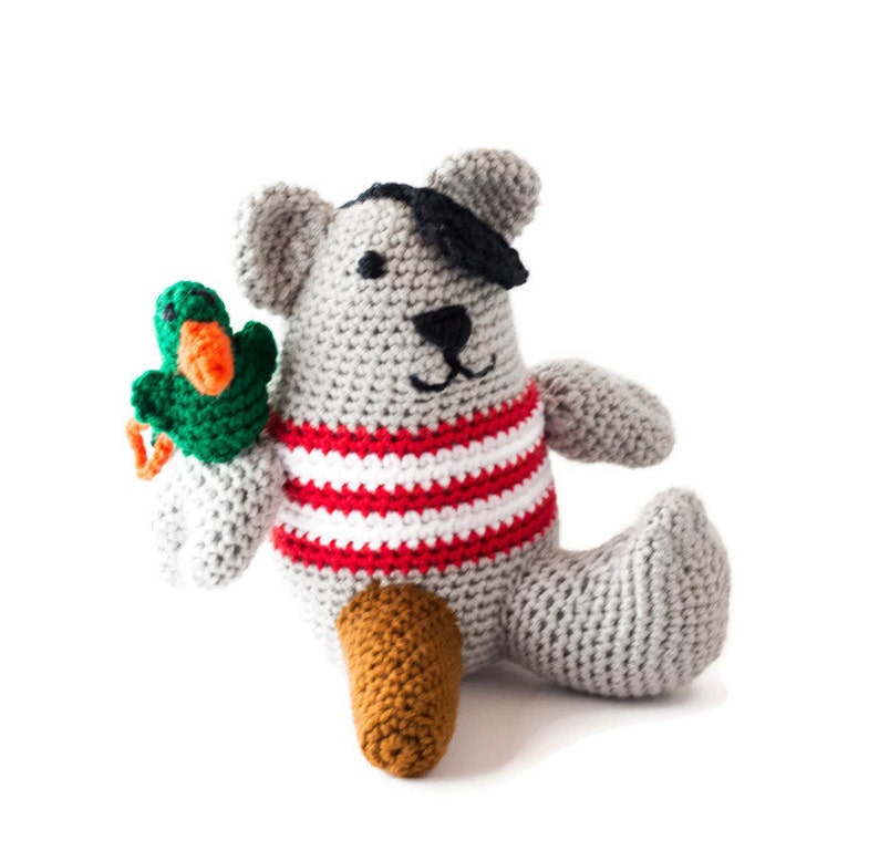 Puede incluir: Un oso de peluche gris tejido a crochet que lleva una camisa a rayas rojas y blancas y un parche negro en el ojo. El oso sostiene un pato verde tejido a crochet en su pata izquierda.