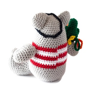 Puede incluir: Un animal de peluche gris tejido a crochet con una camisa a rayas rojas y blancas. El animal tiene un parche en el ojo negro y sostiene un loro tejido a crochet verde y amarillo.