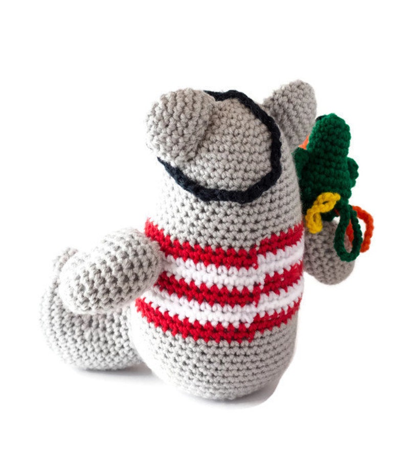 Puede incluir: Un animal de peluche gris tejido a crochet con una camisa a rayas rojas y blancas. El animal tiene un parche en el ojo negro y sostiene un loro tejido a crochet verde y amarillo.