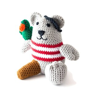 Puede incluir: Un oso de peluche gris tejido a crochet con una camisa a rayas rojas y blancas y un parche negro en el ojo. El oso sostiene un loro verde tejido a crochet en su pata izquierda.
