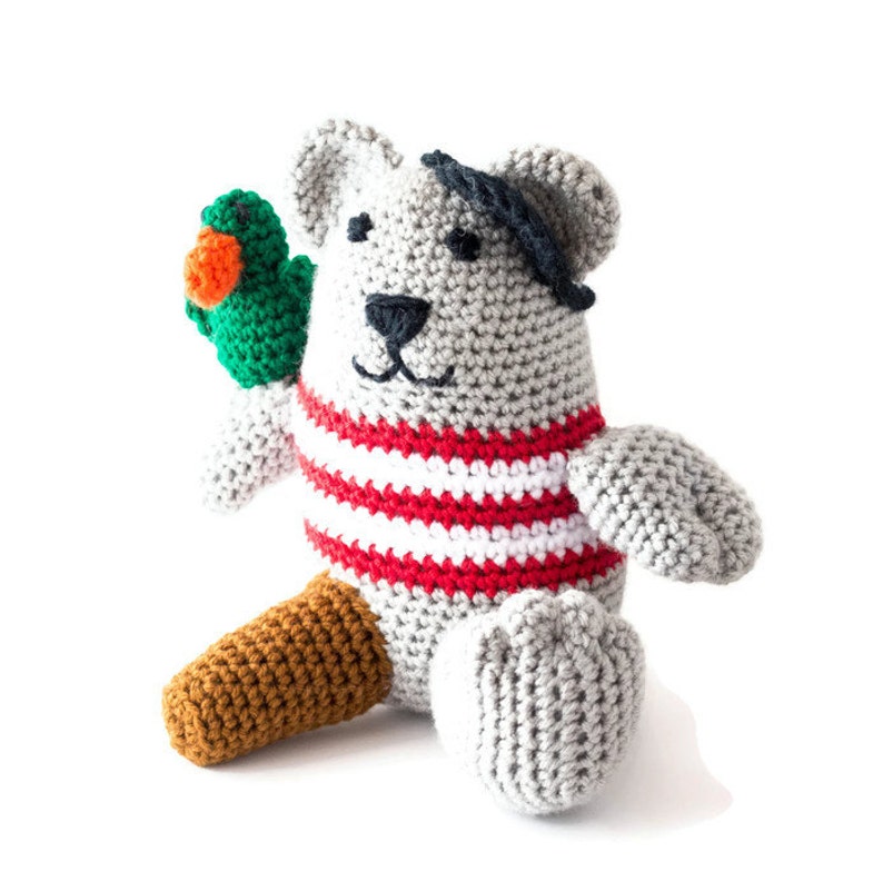 Puede incluir: Un oso de peluche gris tejido a crochet con una camisa a rayas rojas y blancas y un parche negro en el ojo. El oso sostiene un loro verde tejido a crochet en su pata izquierda.