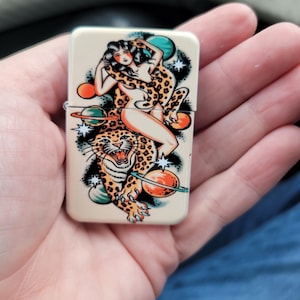 May include: A beige lighter with a colorful tattoo-style design. The design features a woman riding a leopard, surrounded by planets and stars. The leopard is roaring, and the woman is wearing a leopard print. The lighter is held in a hand.