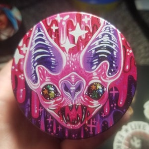 May include: A round metal grinder with a pink, purple, and black design. The design features a cartoon bat with glowing eyes and a dripping effect.