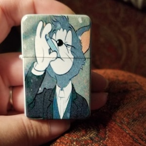 May include: A silver lighter with a cartoon image of Tom from Tom and Jerry. Tom is wearing a suit and tie and has a cigarette in his mouth. The background is a blue and white galaxy print.