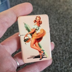 May include: A vintage-style lighter with a colorful image of a woman in a yellow dress holding a basket of green items and a long green object. The lighter has a pink background.