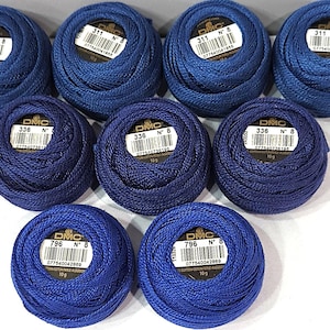 May include: Nine balls of DMC Perle cotton thread arranged in three rows. The top row features four balls of color 311, the middle row has three balls of color 336, and the bottom row has two balls of color 796. Each ball is size 8 and 10 grams.