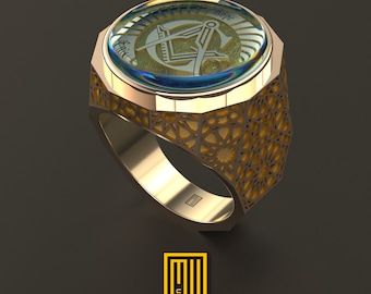 Könnte beinhalten: Ein goldener Ring mit einem detaillierten geometrischen Design. Der Ring hat einen runden, klaren Stein mit einem goldenen Freimaurer-Symbol im Inneren.