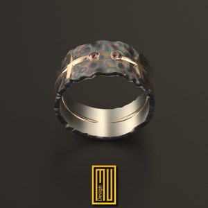 May include: A textured ring with a dark, iridescent finish. The ring features two small, red gemstones and gold-colored accents. The design includes a gold band and a textured outer surface. The ring is a piece of jewelry.