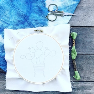 May include: A white embroidery hoop with a pencil drawing of a potted cactus plant. Green embroidery floss is on the right side of the image.